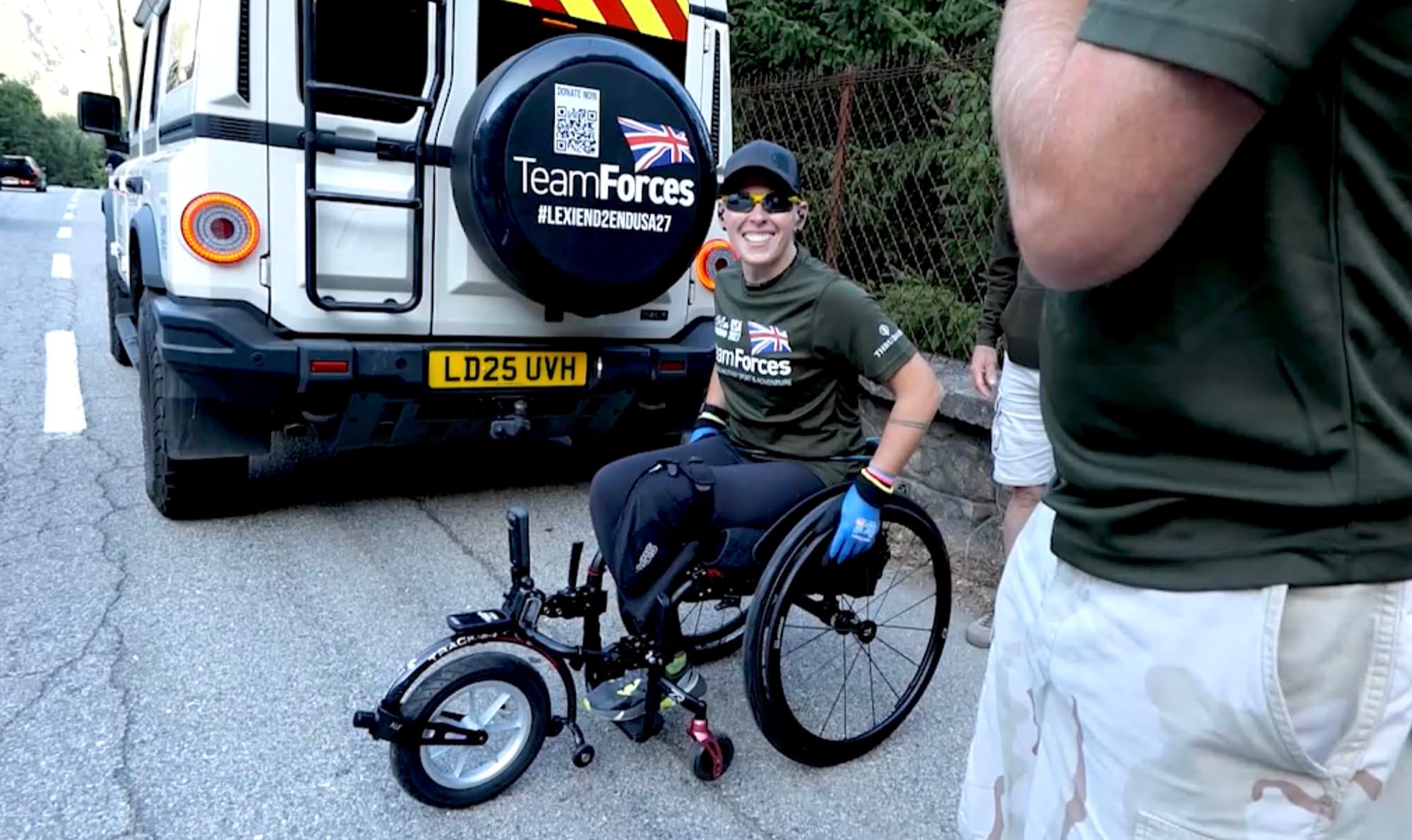 Lexi Chambers smiling in her wheelchair on the road at Alpe d'Huez, beside the TeamForces support vehicle with its #LEXIEND2ENDUSA27 spare-wheel cover.